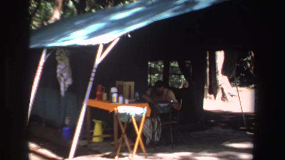 1969 South Africa 8mm frame showing a camp table, yellow pitcher, blue bucket, and a man seated in deep shade.