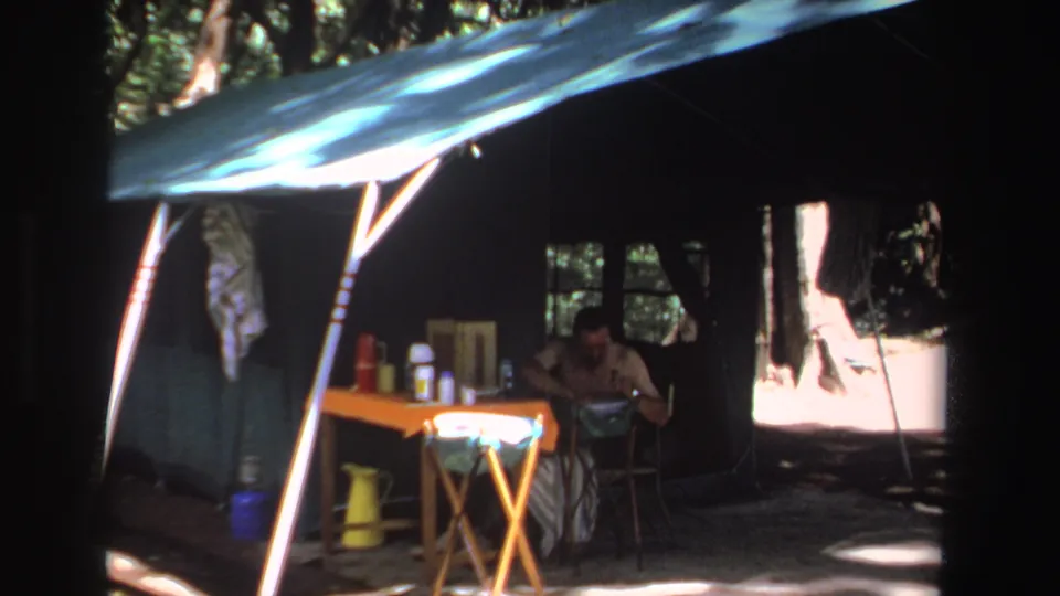 1969 South Africa home-movie frame of a tarp-covered campsite, with a man seated beside a table of camp supplies.