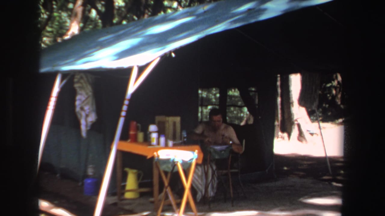 1969 South Africa 8mm frame showing a man seated at an orange table beneath a green tent canopy in wooded shade.