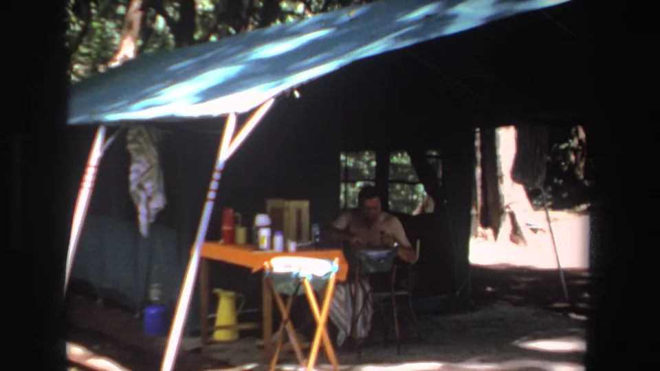 1969 South Africa 8mm frame showing a man seated at an orange table beneath a green tent canopy in wooded shade.