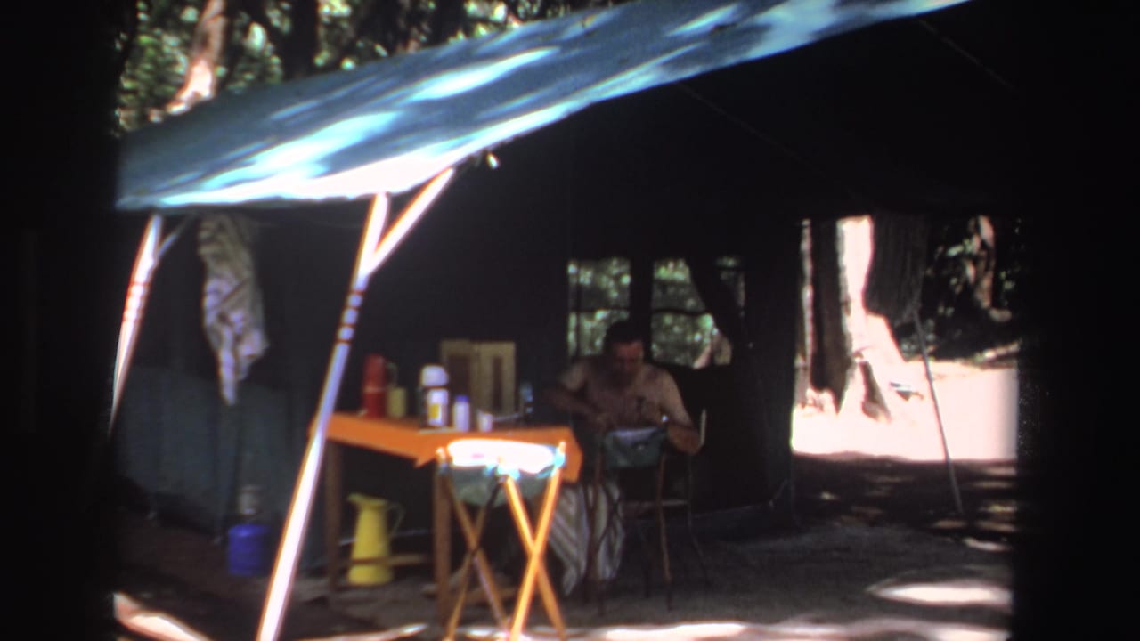 Seated man at an orange table under a tent canopy with camping containers in shade.