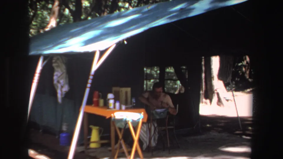1969 South Africa home-movie frame showing a shaded campsite with a man, orange folding table, yellow pitcher, and trees.