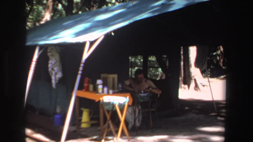 1969 South Africa 8mm frame capturing a man at a shaded campsite table beneath a green canopy, surrounded by trees.