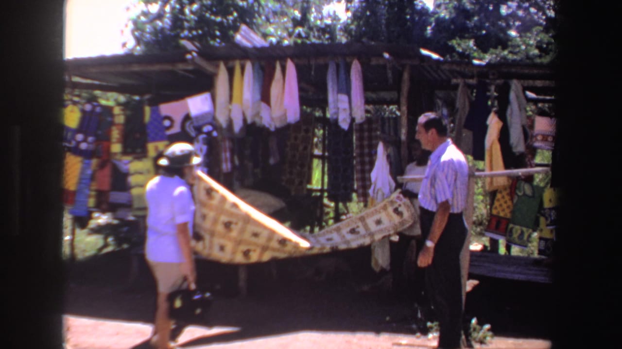 1969 South Africa home-movie frame of a woman in a hat and a man viewing patterned fabric at an outdoor market stall.