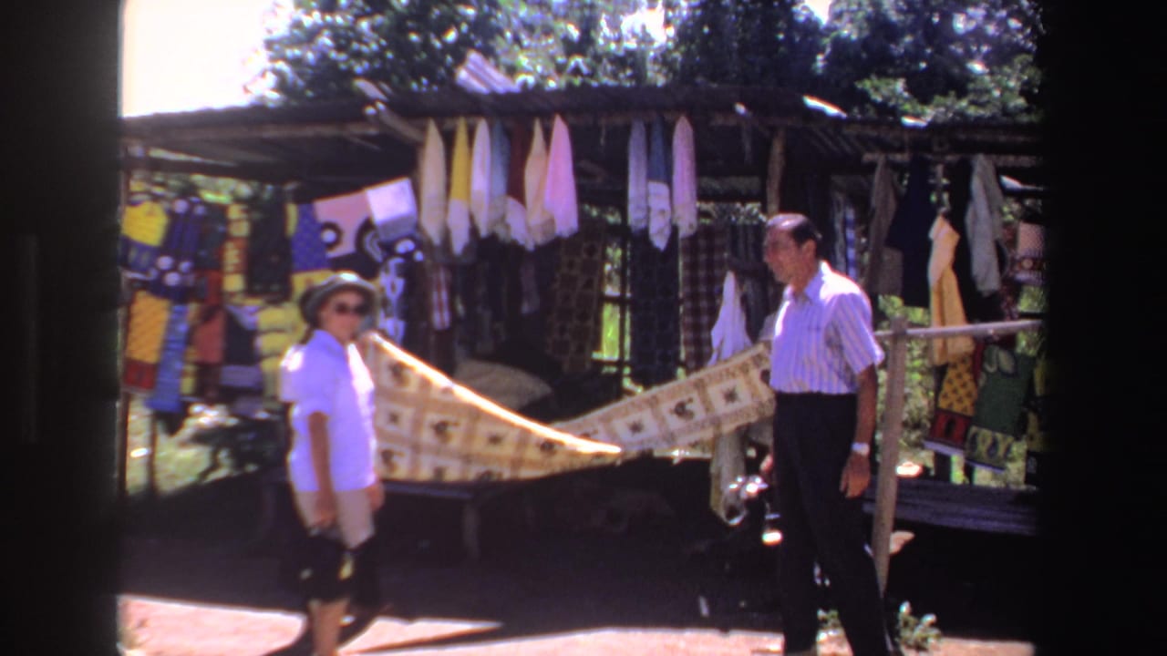 Woman walking past a fabric display while a man stands near a shaded market stall.