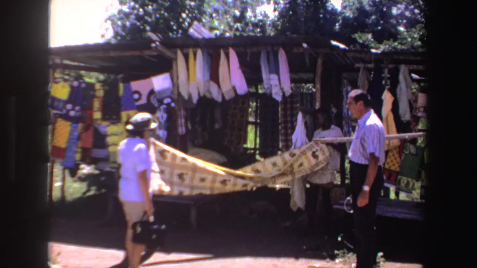 1969 South Africa home-movie frame of two adults beside a long patterned textile under a shaded outdoor stall.