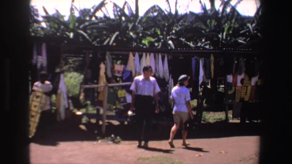 1969 South Africa frame showing the couple leaving a shaded textile stall, with hanging fabrics and large tropical leaves behind.