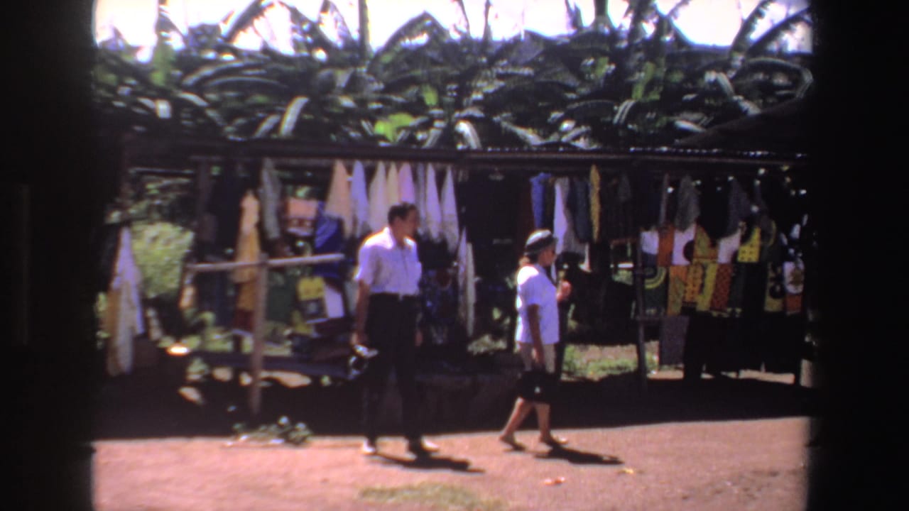 Two shoppers walk past hanging fabrics at an outdoor market stall.