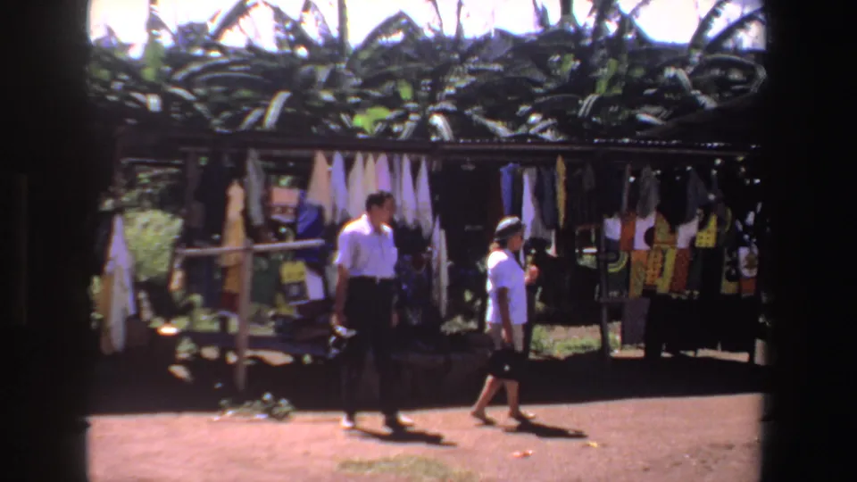 In 1969 South Africa home-movie footage, two shoppers pass fabric racks under a corrugated market roof.