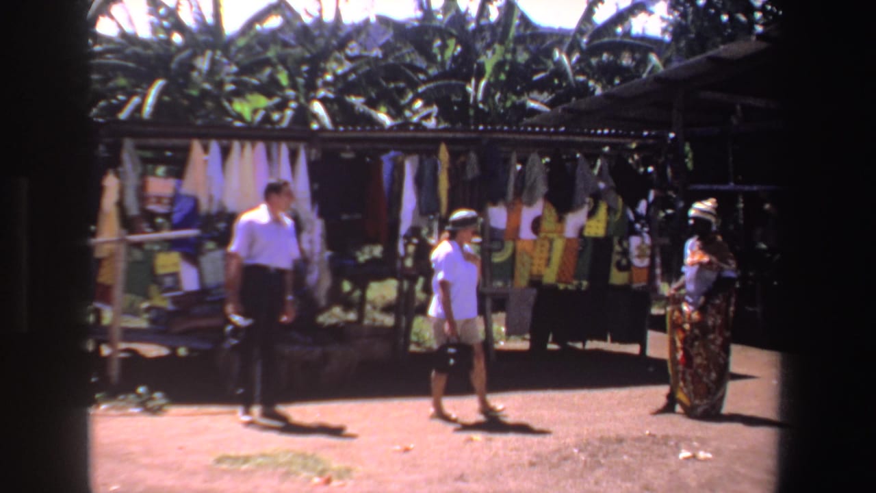 Two shoppers approach a vendor beside colorful hanging textiles in a South African outdoor market scene.