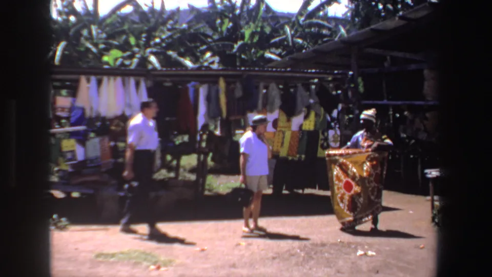 A market vendor displays a vivid patterned cloth while two shoppers stand nearby in the 1969 South Africa footage.