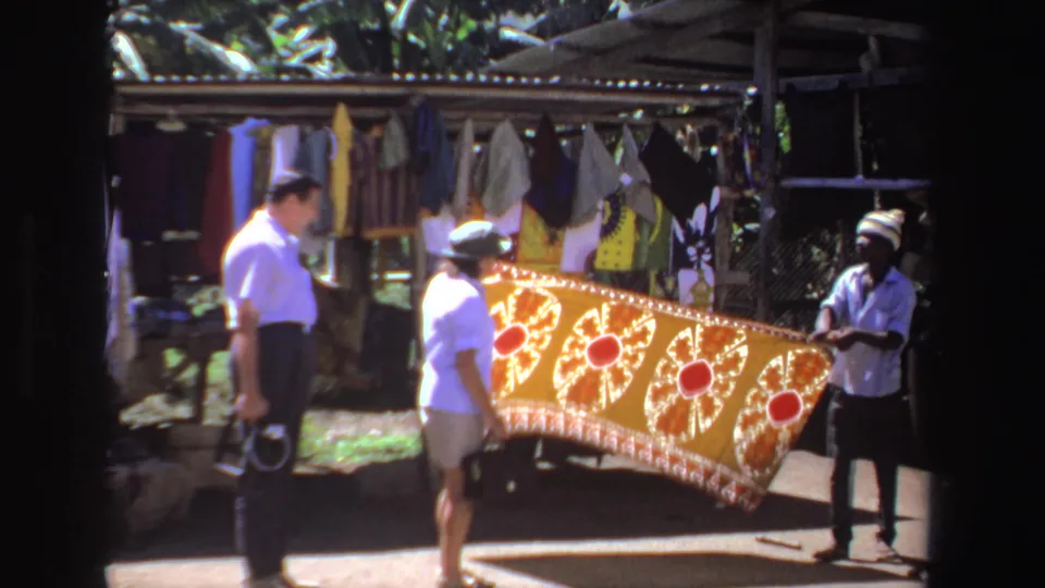 A vendor holds out a large orange patterned textile for shoppers at an outdoor South African market stall.