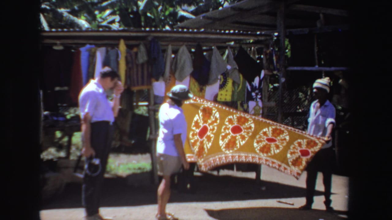 A man and woman face a vendor holding a bright patterned tablecloth in front of hanging textiles.