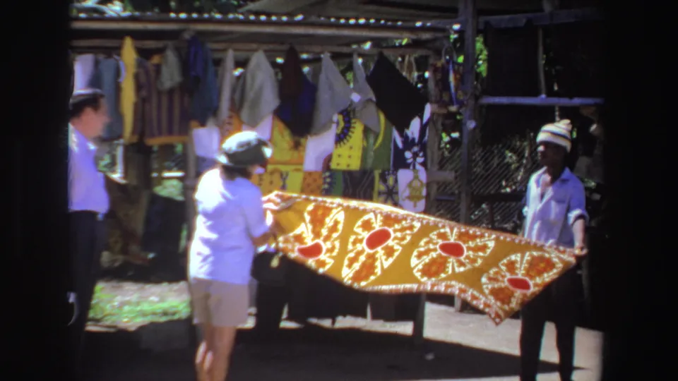 The visitor-shot 1969 market footage shows a woman closely inspecting a red-and-gold tablecloth at a South Africa bazaar.