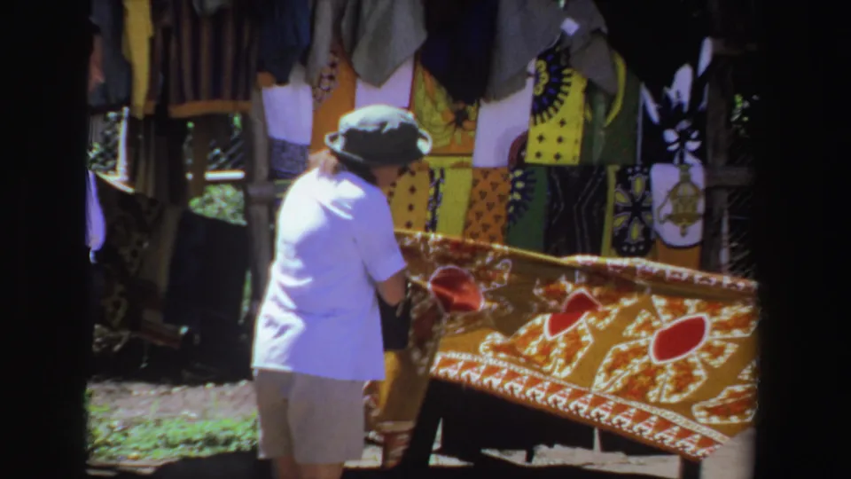 A closer 1969 South Africa frame shows the shopper handling the red-and-gold tablecloth near hanging textiles at the stall.
