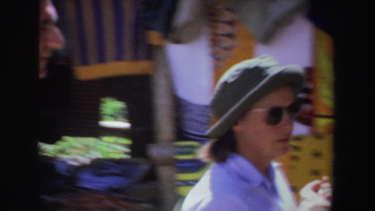 Woman in hat and sunglasses passes hanging garments at a market.
