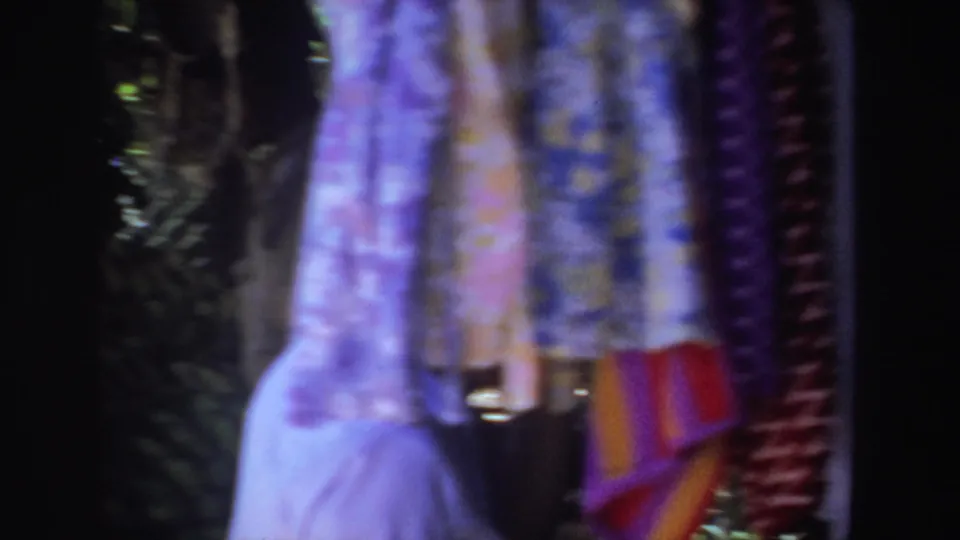 Colorful hanging dresses and textiles dominate the frame at the outdoor market, with only a partial figure visible near the stall.