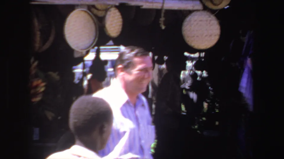 1969 South Africa market frame with a smiling tourist, a foreground figure, and woven craft items hanging in the background.
