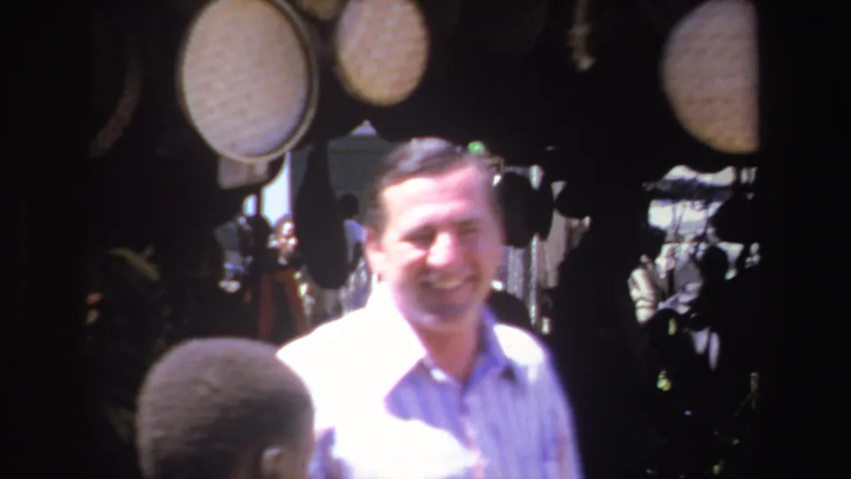 In South Africa, 1969, a smiling man in a pale blue shirt stands near hanging woven baskets in an outdoor market.