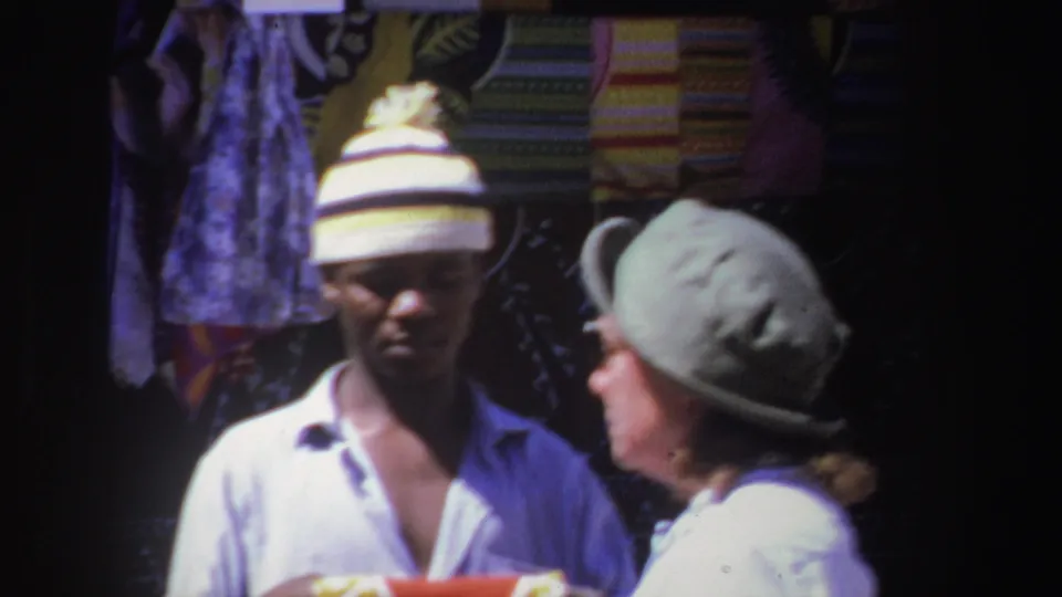 A man in a striped knit hat faces a woman in a green hat beside hanging patterned fabrics at a South African market in 1969.