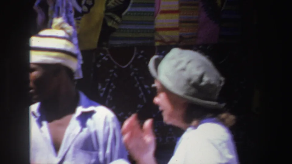 The vendor and a woman in a green hat appear to speak closely in front of patterned textiles at the outdoor market.