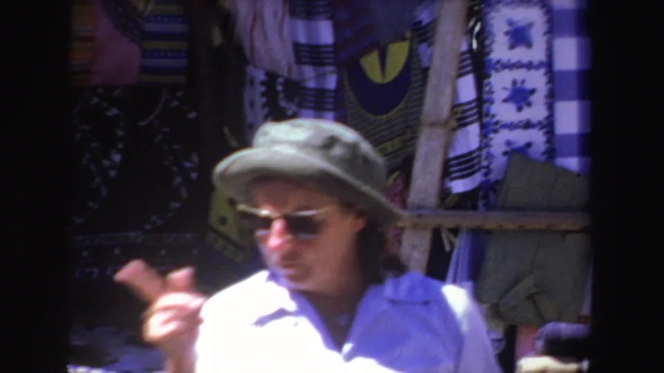 A man wearing sunglasses and a soft hat gestures near hanging patterned cloths in the 1969 South Africa market scene.