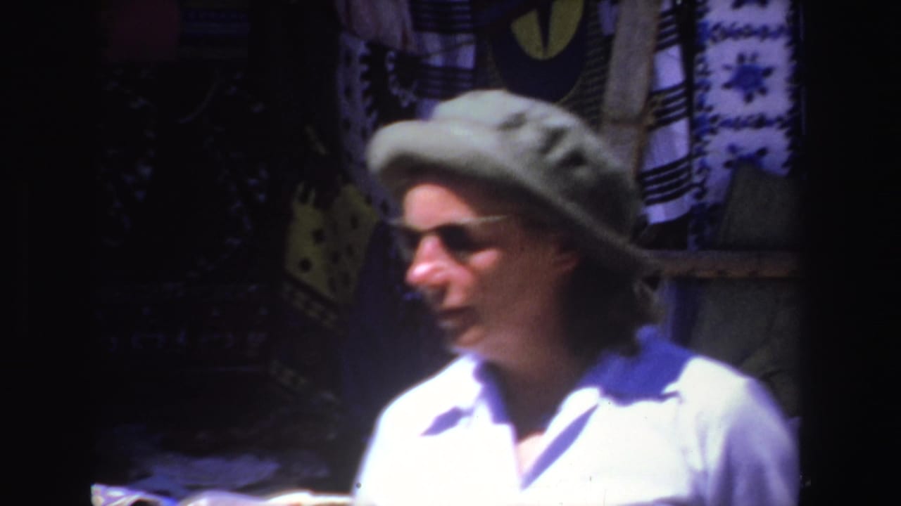 In 1969 South Africa footage, a woman in a green hat and sunglasses is seen close to hanging textiles at an outdoor market stall.