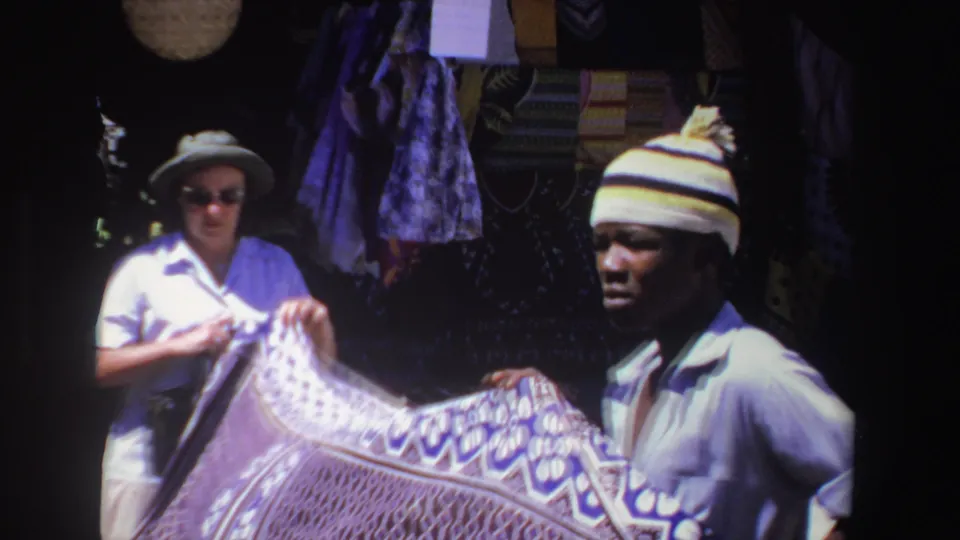 In a South Africa market scene from 1969, a woman in sunglasses and a vendor in a striped cap hold a patterned textile.