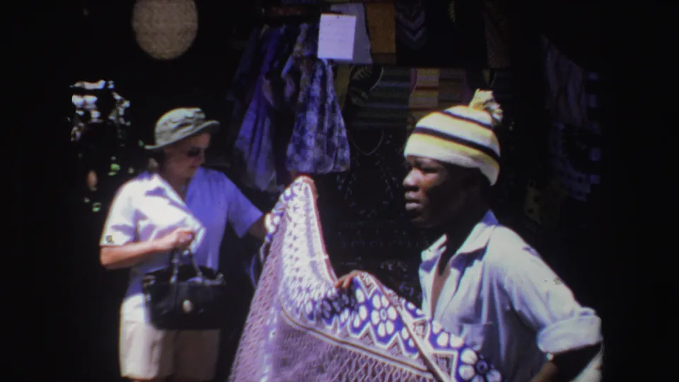 The 1969 South Africa home-movie frame shows a vendor holding a patterned textile as a woman stands nearby at the market stall.