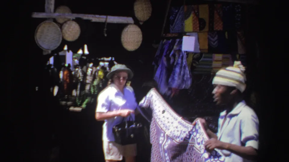 A 1969 South Africa market frame shows a woman and vendor facing each other across a patterned textile under hanging woven goods.