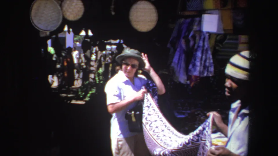1969 South Africa 8mm frame showing an adult tourist in a sun hat examining a patterned textile at an open-air market stall.