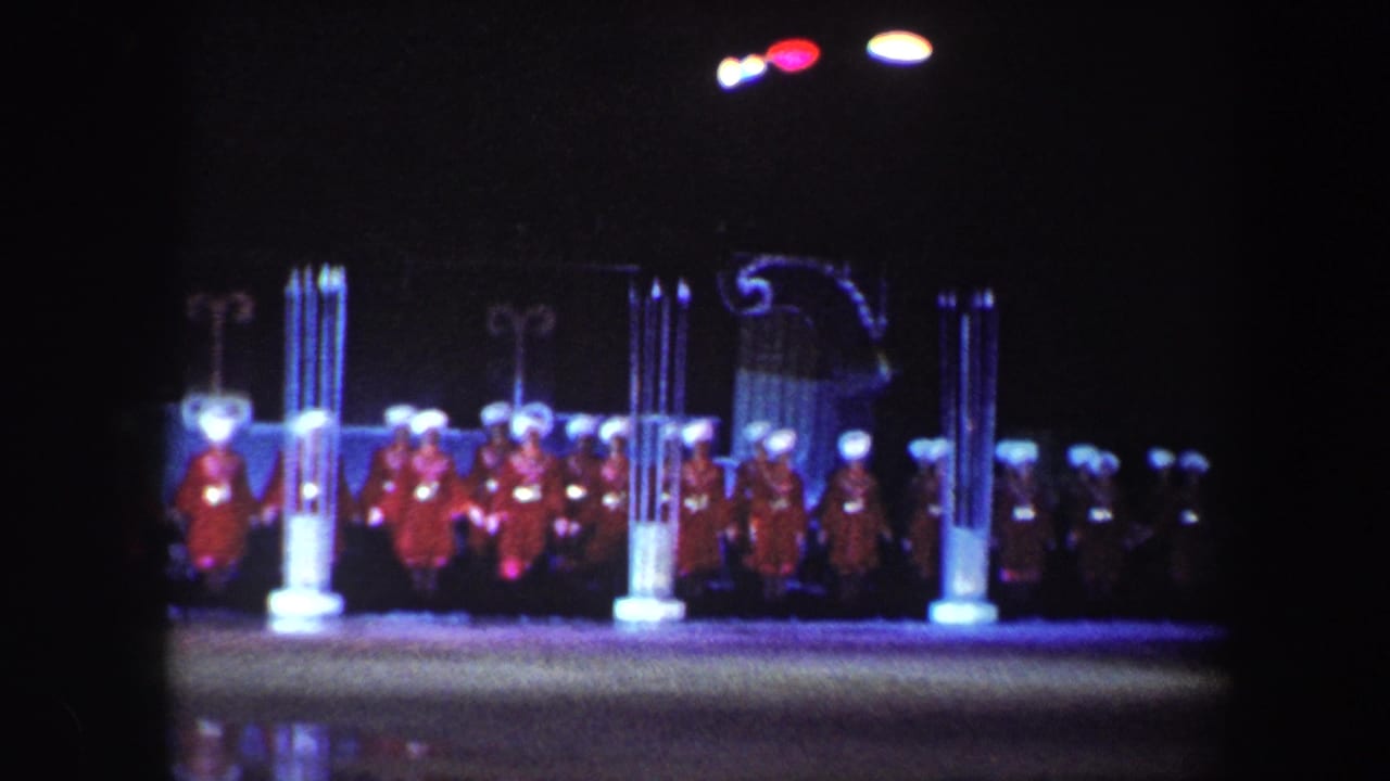 Red-costumed performers stand on a dark stage between illuminated column props.