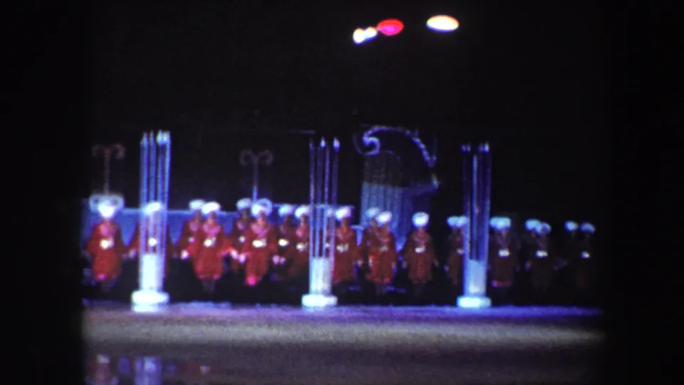 1960 Baltimore home-movie frame showing a distant stage group in red costumes and white hats under colored lights.
