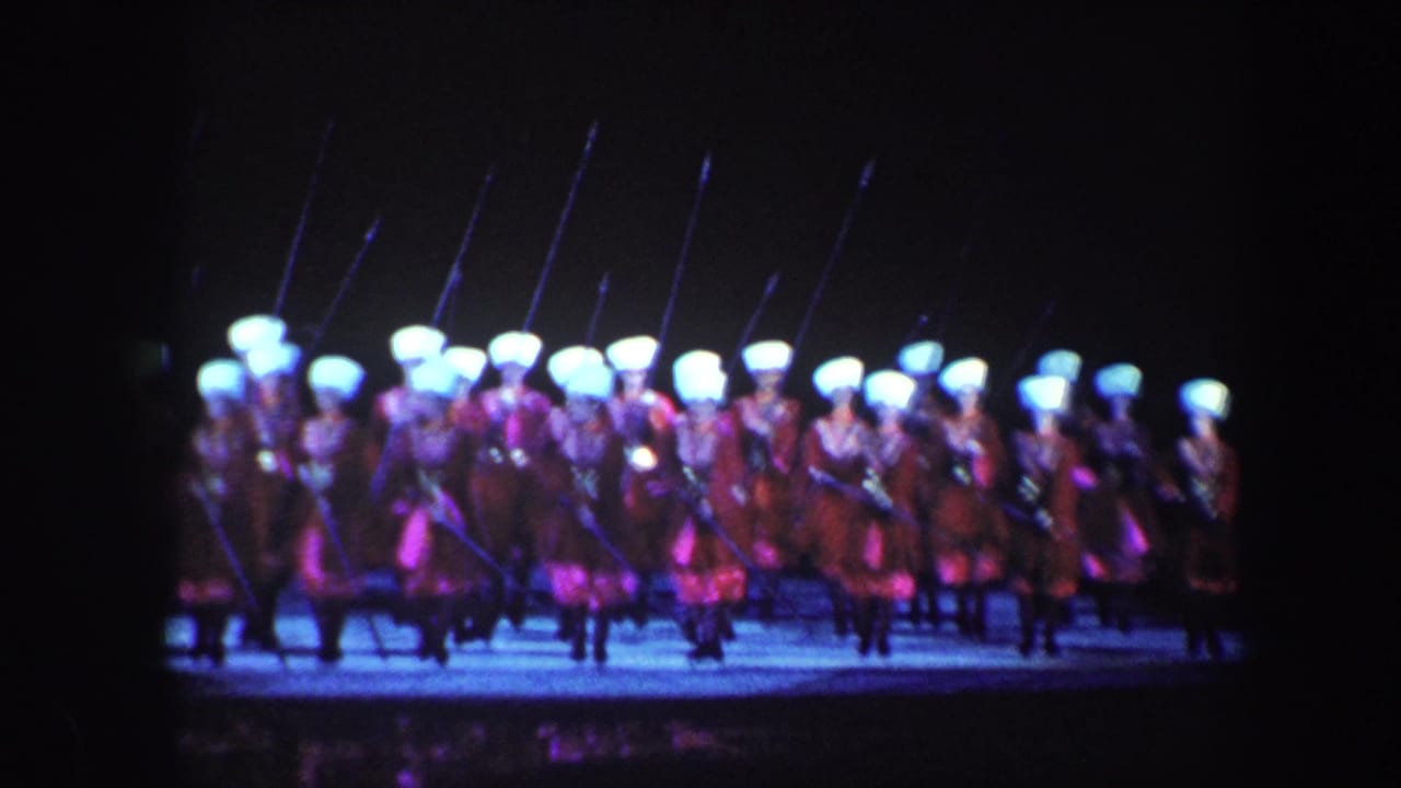 A line of performers in red costumes carry long pikes across a lit stage.