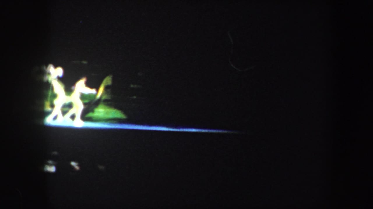 Small figures perform under green and white stage light on a dark stage.