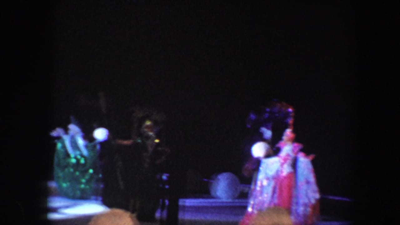 Costumed circus performers and clowns stand under spotlights on a dark stage.
