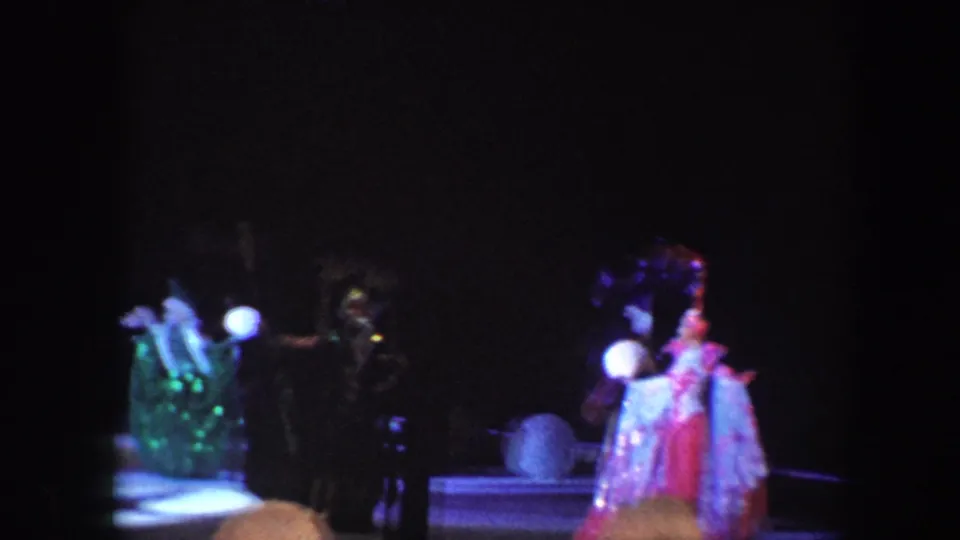 In 1960 Baltimore home-movie footage, clowns and costumed performers appear on a dim circus stage under bright spotlights.