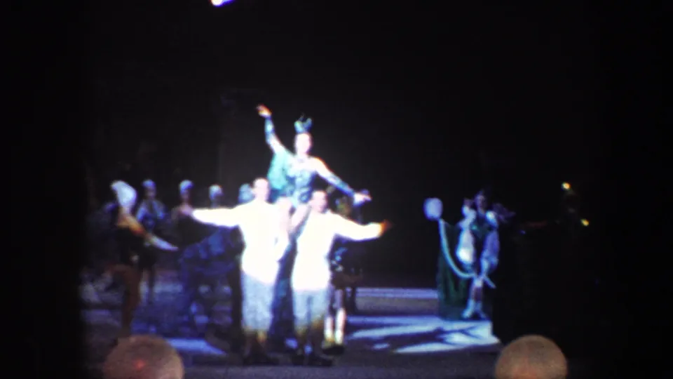 A staged circus act shows an acrobat raised above two performers, with additional costumed figures visible in the background.