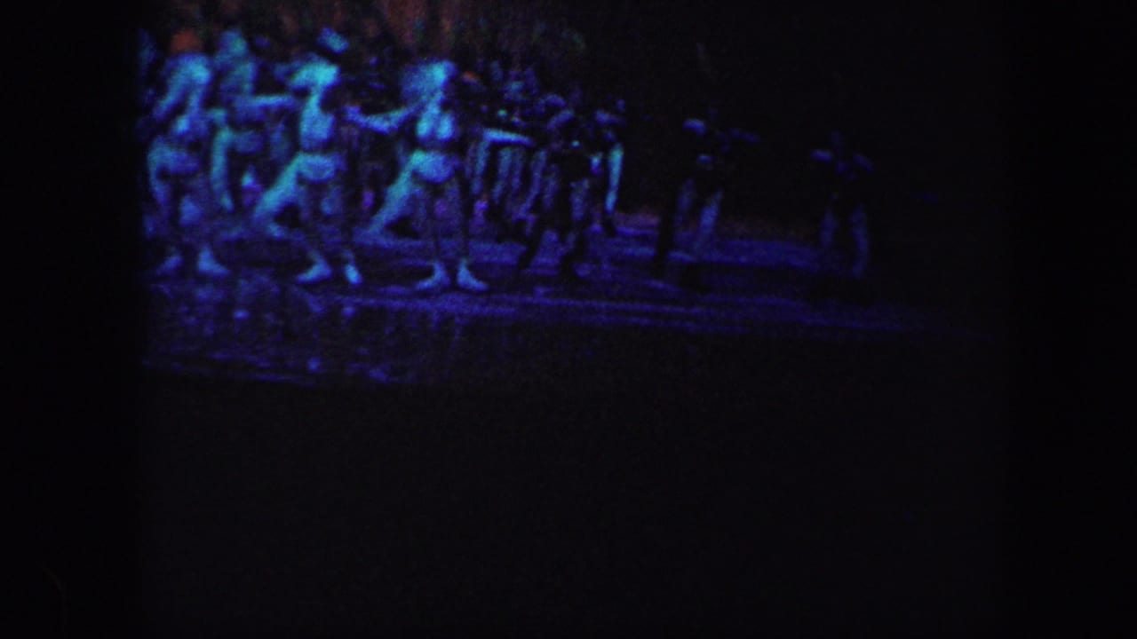 In 1960 Baltimore home-movie footage, distant performers skate in formation under blue-purple theatrical lighting on a dark ice rink.