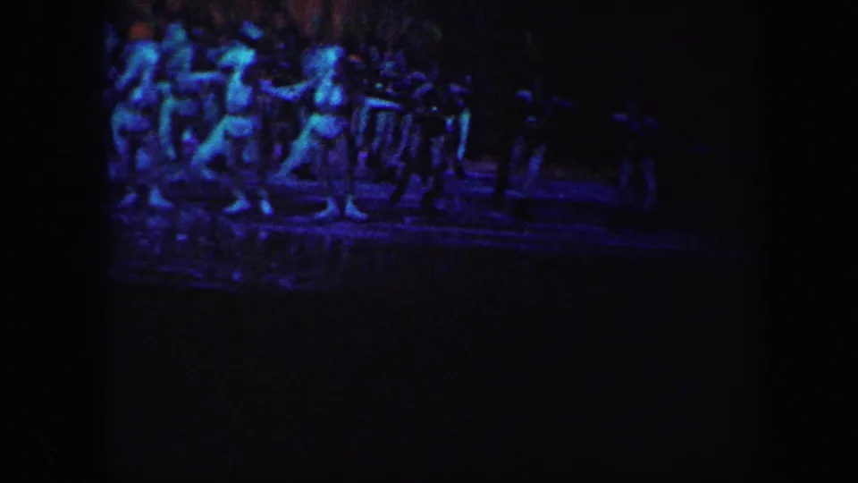 In 1960 Baltimore home-movie footage, distant performers skate in formation under blue-purple theatrical lighting on a dark ice rink.