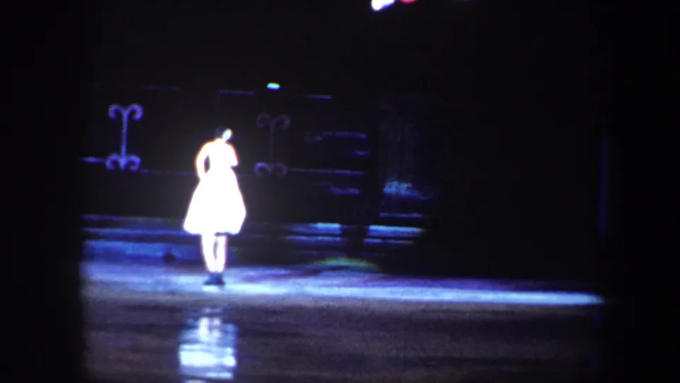 A solo skater in a white costume stands in a spotlight on the ice, with set pieces visible in dark 1960 Baltimore home-movie footage.