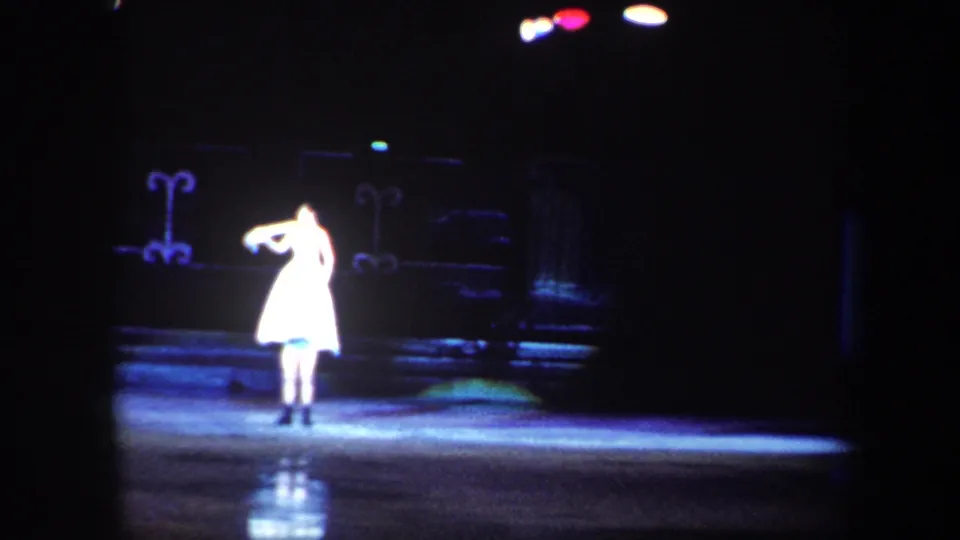 The solo skater poses in a bright white costume under a spotlight, with dark theatrical scenery behind her in the 1960 Baltimore clip.