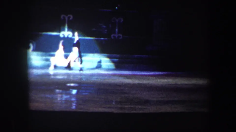 A circa-1960 Baltimore home-movie frame shows skaters under bright stage light on a dark ice rink.