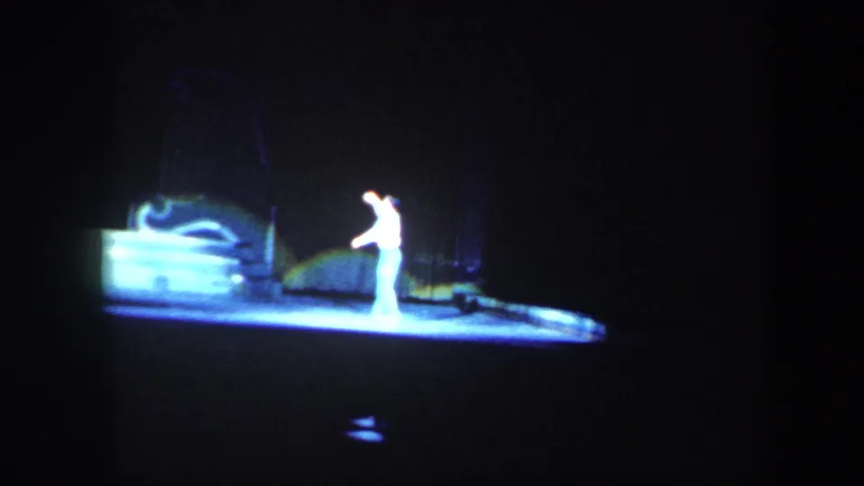 A single white-costumed skater is isolated by a spotlight on the dark rink in the 1960 Baltimore footage.