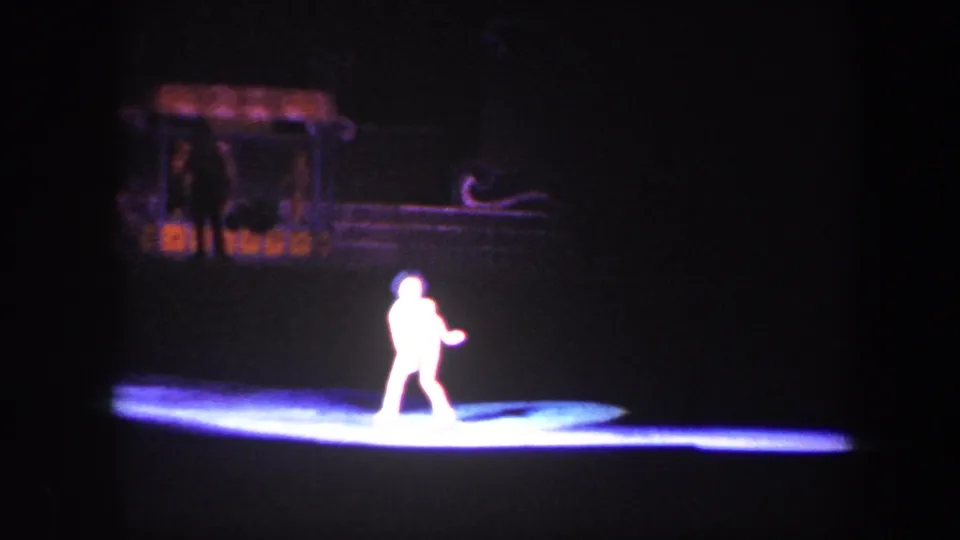 A solo skater crosses the illuminated rink while darker theatrical scenery remains in the background.