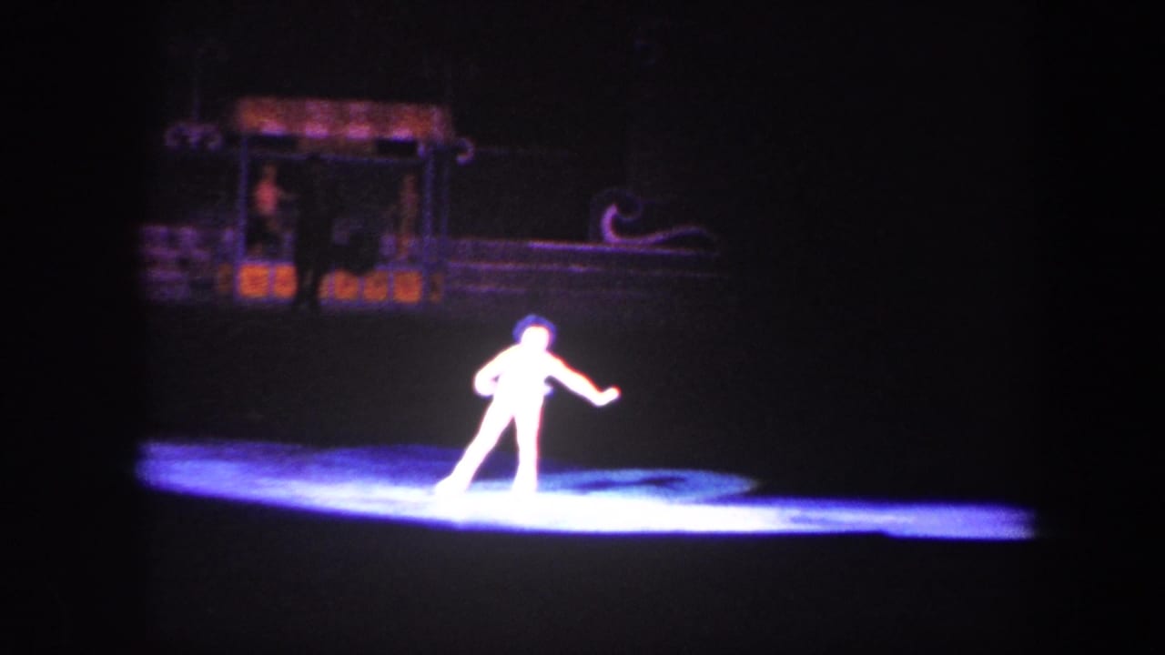 Lone skater in a bright spotlight on a dark ice rink with set pieces behind.