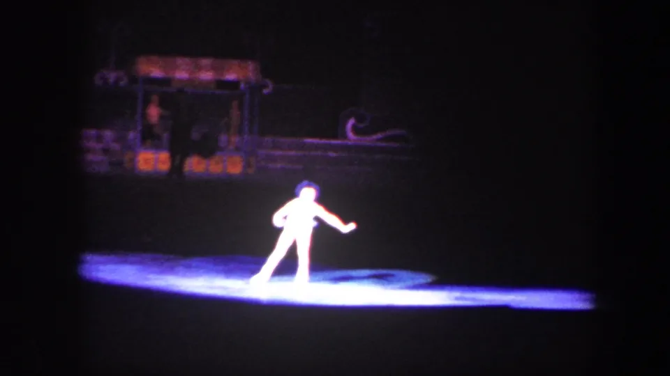 In supported 1960 Baltimore home-movie footage, a single skater performs under a white-blue spotlight on a dark rink stage.
