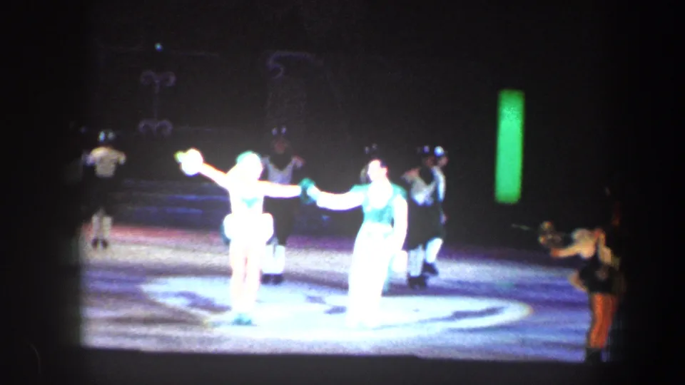 Two performers are strongly overexposed by stage lights as other costumed skaters remain visible in the background.