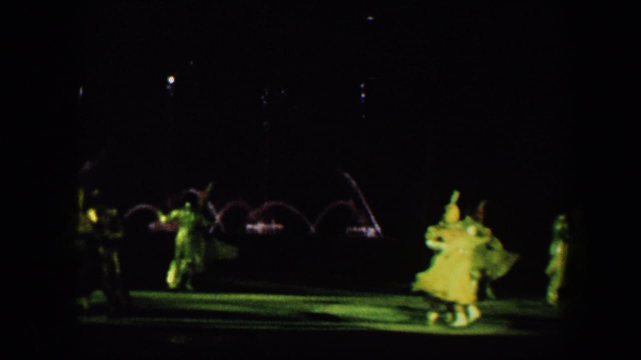 Baltimore 1960 home-movie frame showing costumed ice skaters on a dark rink with green theatrical light and fountain arcs behind them.