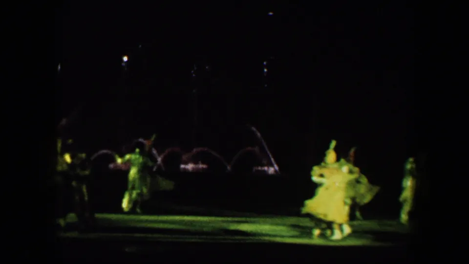 Baltimore 1960 home-movie frame showing costumed ice skaters on a dark rink with green theatrical light and fountain arcs behind them.